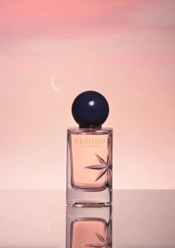 Flacon de parfum “BEYOND STARS” avec bouchon sphérique foncé, sur fond dégradé rose.